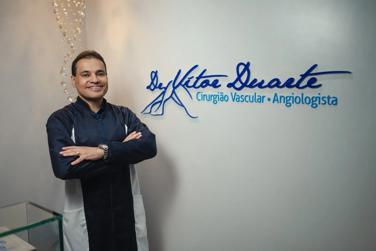 Dr. Vitor Duarte de jaleco em seu consultório, ao lado da logomarca Cirurgião Vascular e Angiologista
